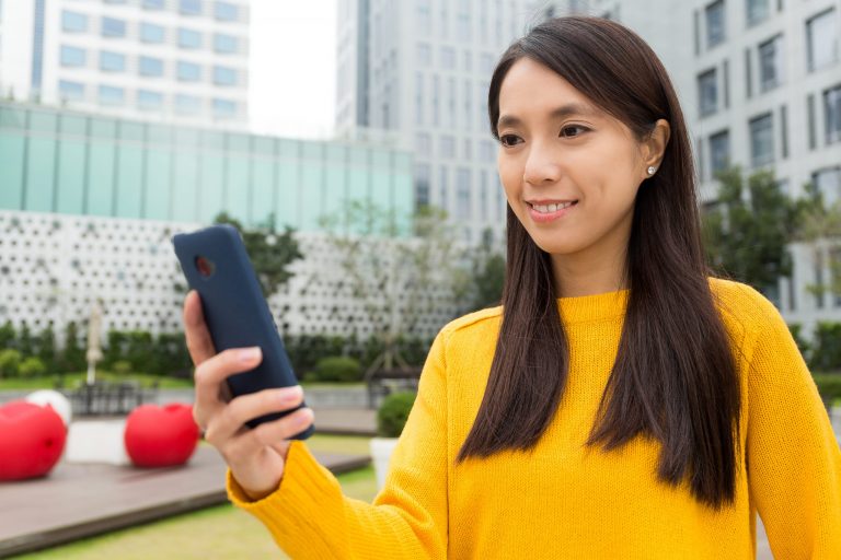 Smiling woman using smartphone outdoors, representing Mocean SMS messaging campaign web portal for easy customer communication.
